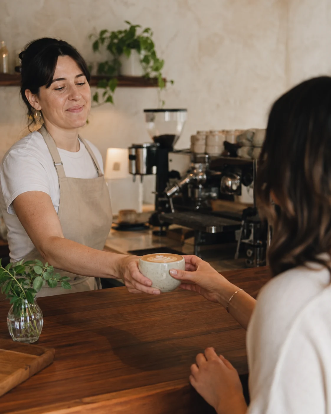 Barista handing a ceramic flat white to an adult guest across a walnut café counter.