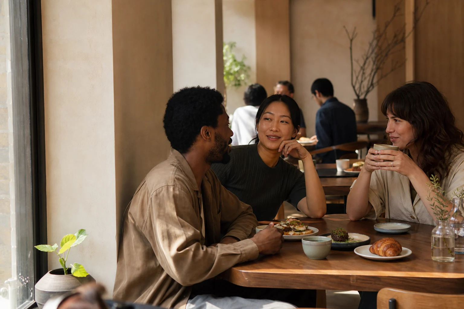 Three adult friends talking over coffee, pastries and brunch plates in a warm café.